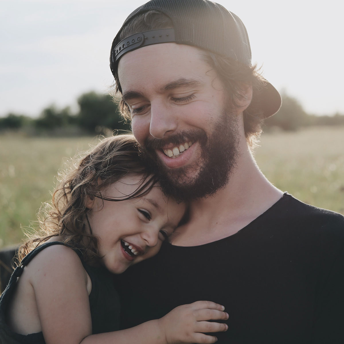 Father holding daughter in his arms. Both are smiling and the sun is shining on the foliage in the background.