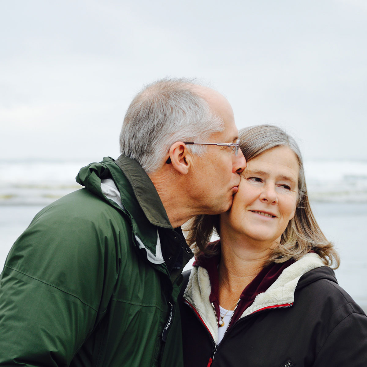 Older couple in walking clothes at the beach. Man is giving woman kiss on the cheek.