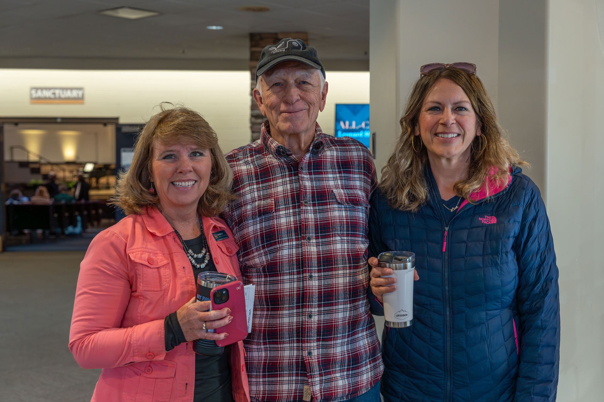 Three adults smiling indoors holding travel mugs.
