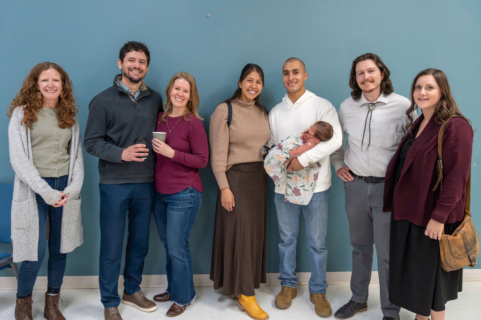 Seven adults posing indoors, one holding newborn baby