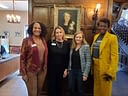 Four women pose together beneath a framed portrait