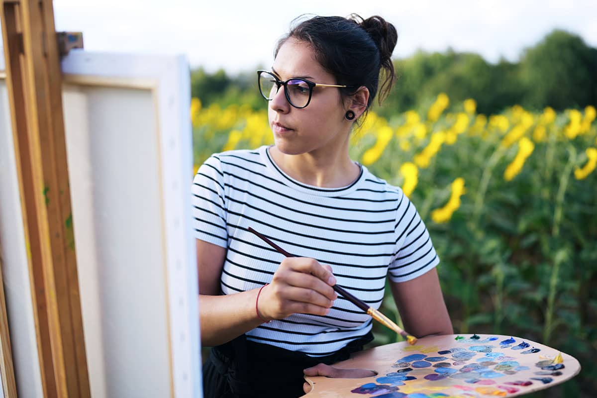 High school student painting on an easel outdoors in a field of sunflowers