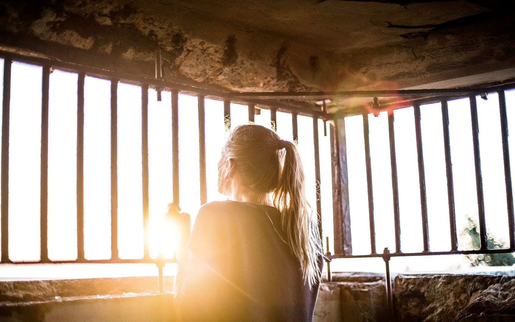 A young woman in a cell clutches bars and peers towards the sunrise