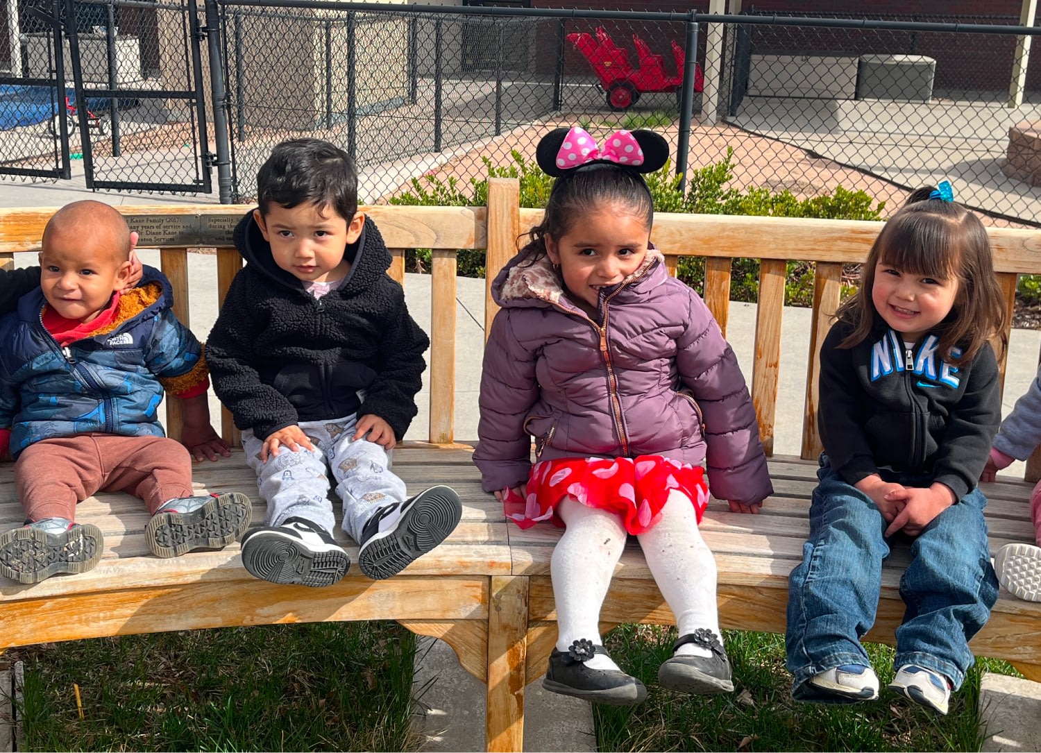 Four children sitting on a wooden bench outdoors.