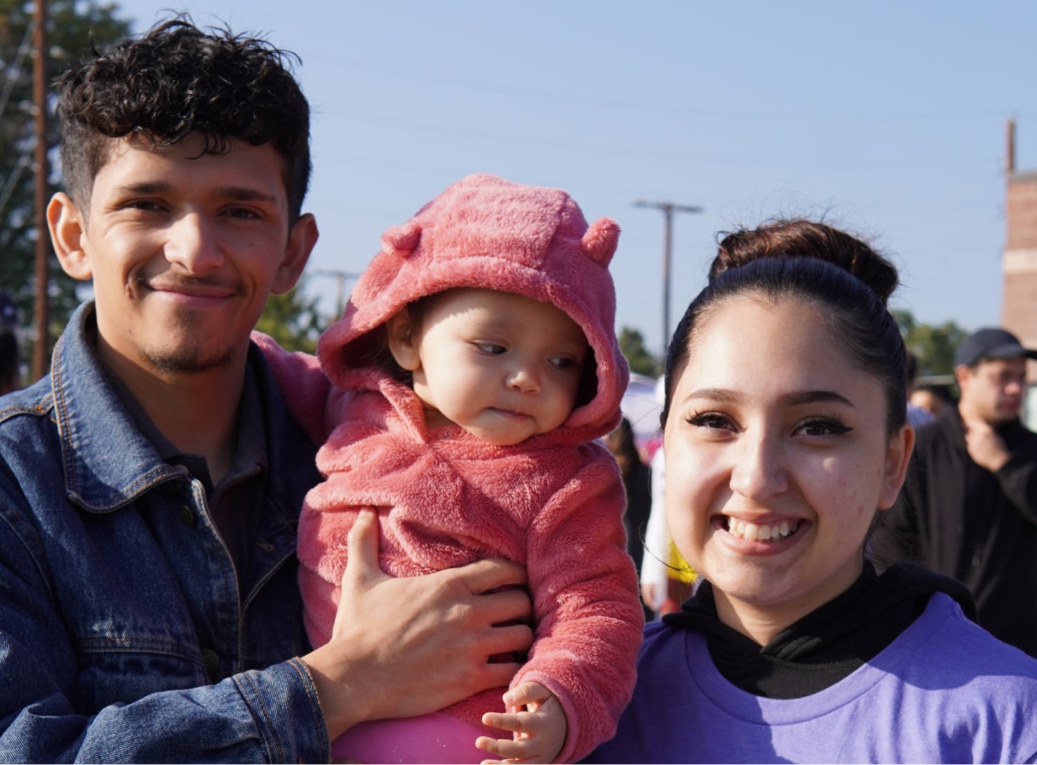 Smiling family with baby in pink hoodie outdoors.