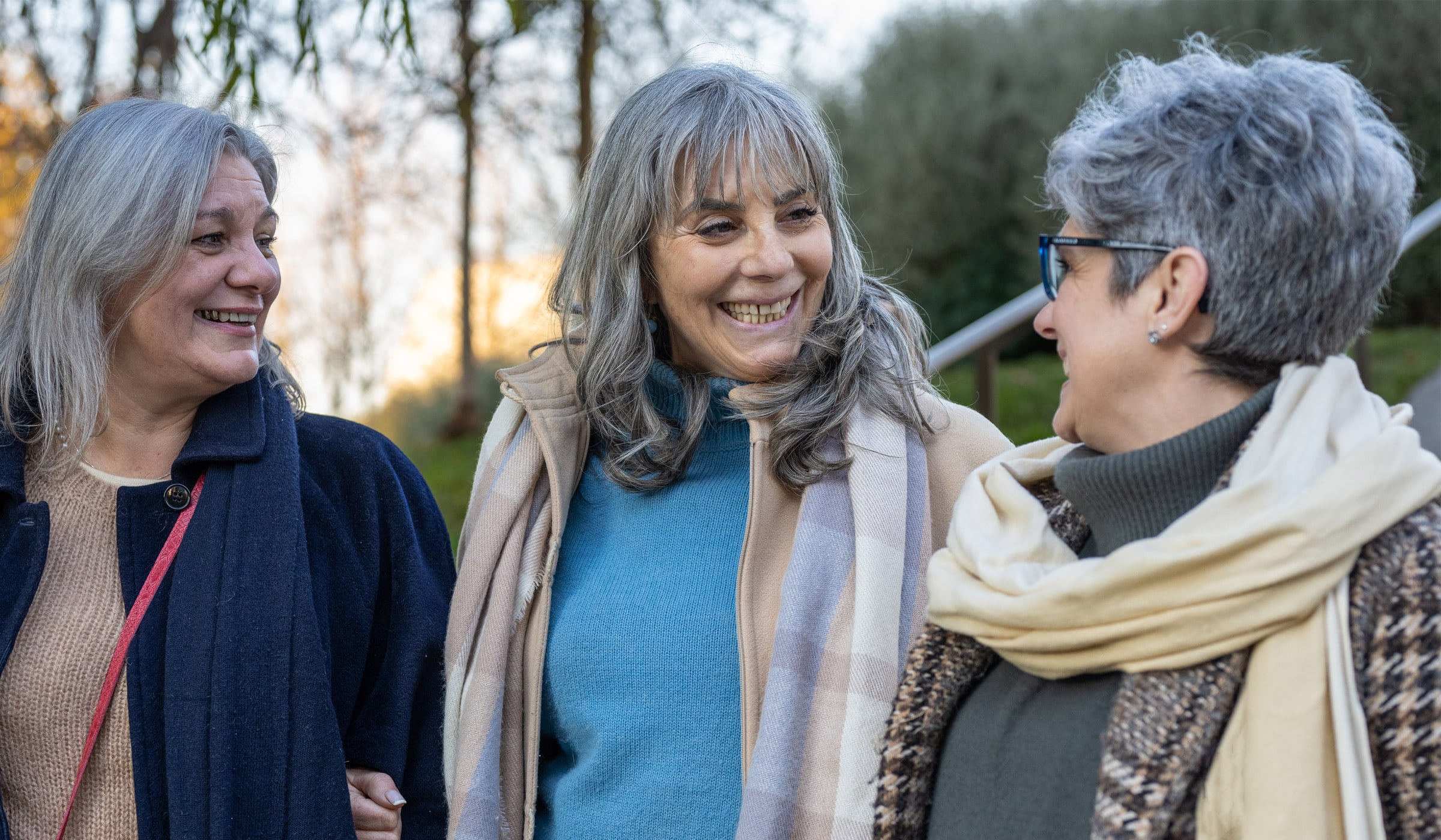 Three women smiling outdoors in fall scenery.