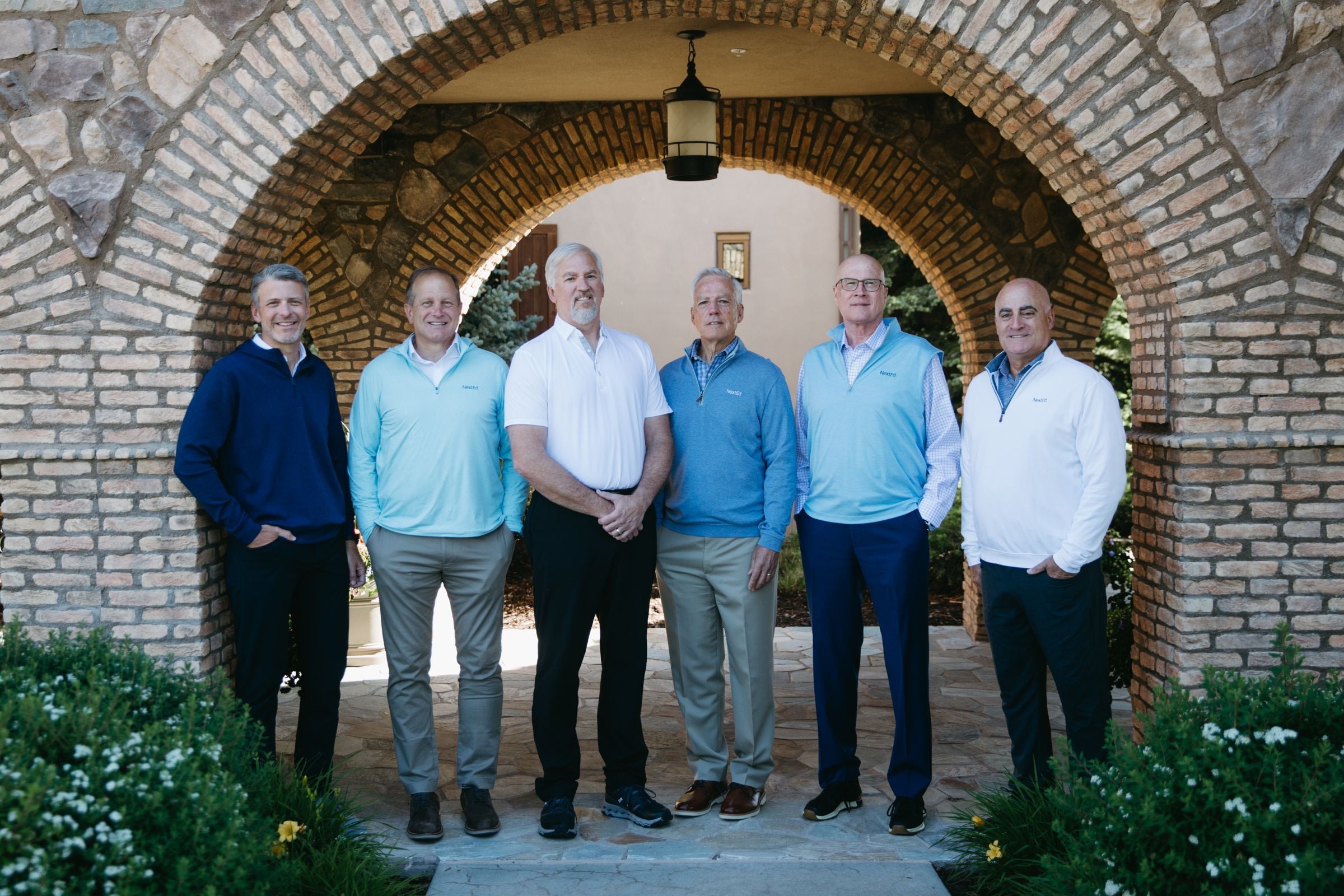A group of six men standing together under a brick archway, posing for a photo. They are dressed in smart-casual attire, mostly wearing polo shirts and pullovers. The setting appears to be outdoors in a landscaped area with stone paving, greenery, and a building visible in the background.