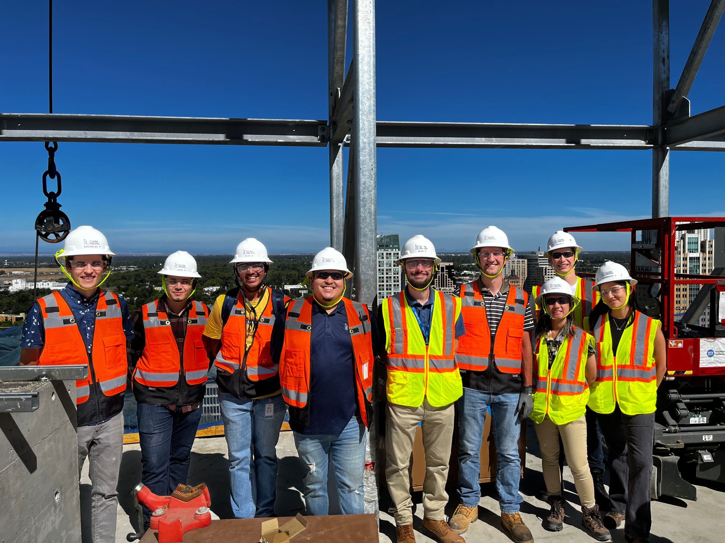 A group of nine people wearing hard hats, safety vests, and work boots standing on a construction site high above the city, with steel beams and equipment around them and a cityscape in the background.