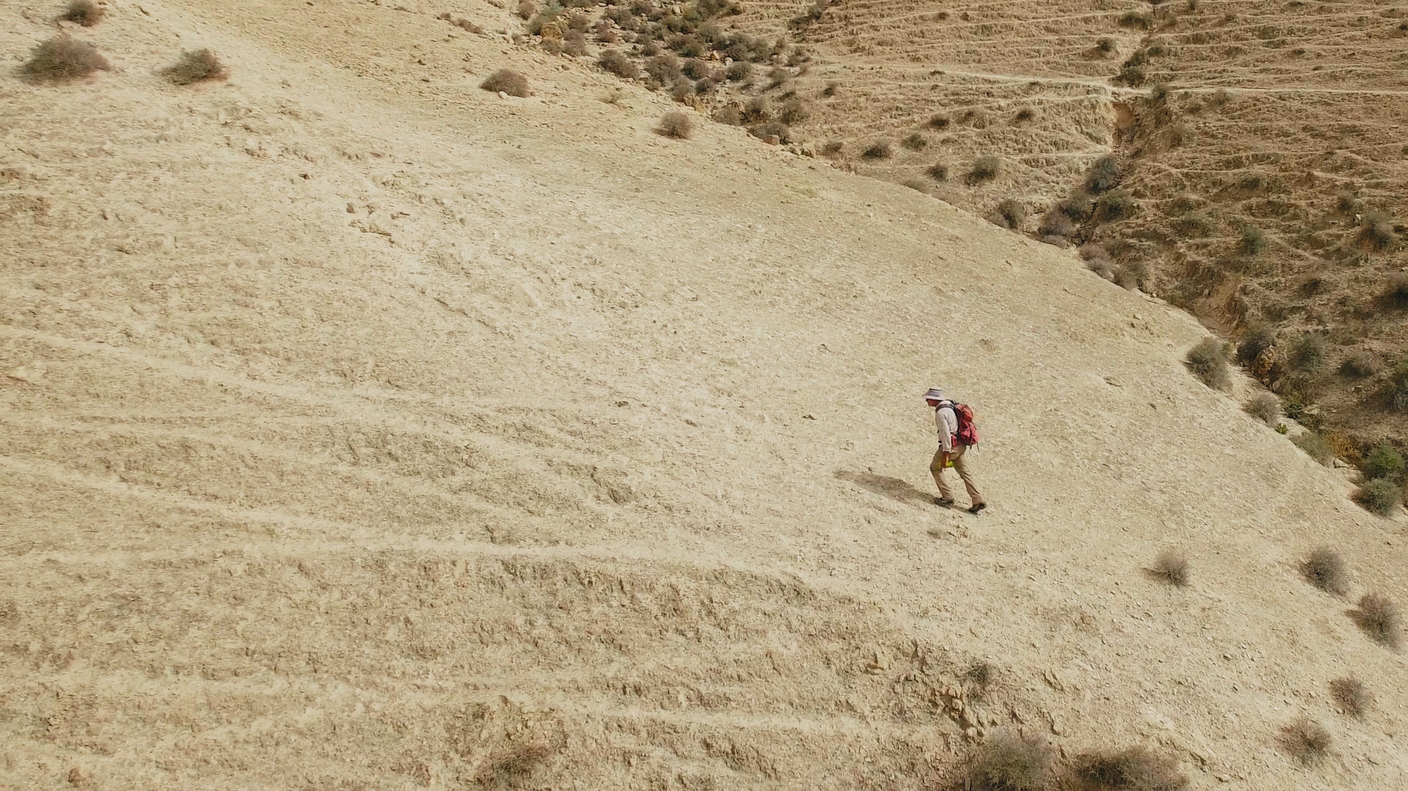 Hiker climbs arid hillside with red backpack.