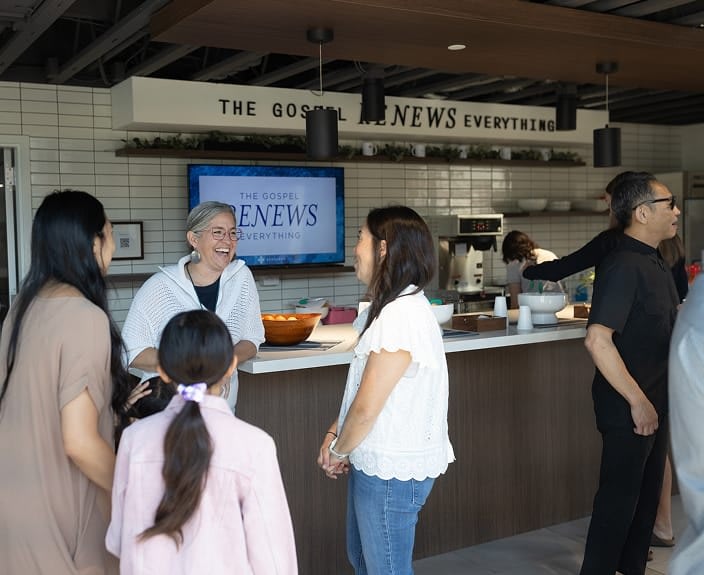 A diverse group of church members talking and laughing at a coffee bar in a church lobby, with a screen in the background displaying the phrase "The Gospel Renews Everything."
