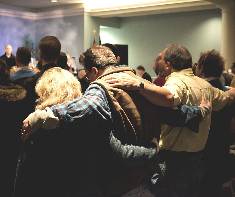 room of praying people from behind two couples arm in arm praising God